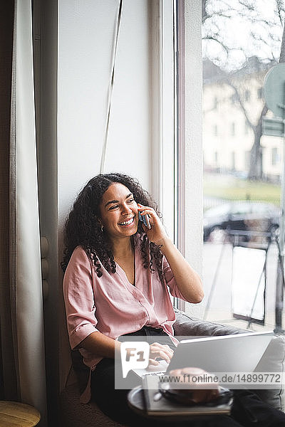 Lächelnde Jungunternehmerin spricht durch ein Smartphone  das im Büro auf dem Fensterbrett sitzt