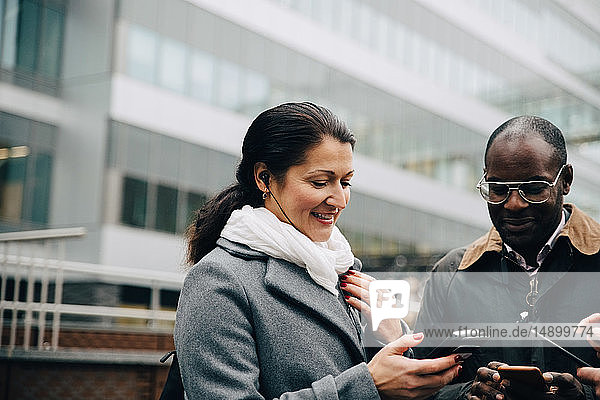 Lächelnde Kollegen  die Mobiltelefone benutzen  während sie gegen Gebäude in der Stadt stehen