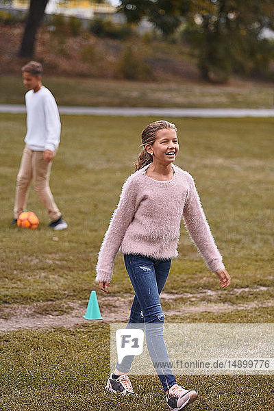 Lächelnde Schwester geht auf Grasfeld  während der Bruder im Hintergrund im Park Fussball spielt