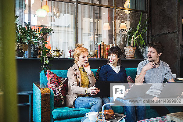 Smiling colleagues looking at female manager sitting with laptop on sofa at creative office