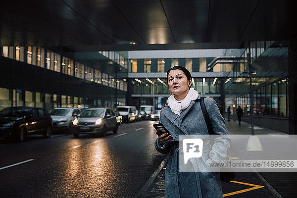 Geschäftsfrau mit Mobiltelefon  die weg schaut  während sie auf der Straße in der Stadt steht