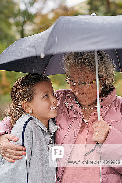 Großmutter umarmt Enkelin  während sie mit ihr im Park während der Regenzeit den Schirm teilt