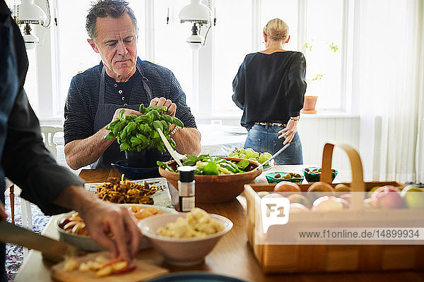 Reife Männer bereiten das Essen vor  während die Freundin zu Hause im Hintergrund steht