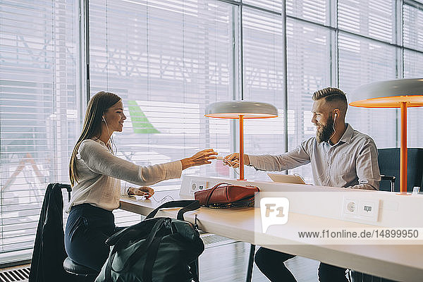 Smiling businessman giving charger to young businesswoman while sitting at airport