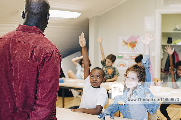 Schüler heben die Hand  während sie den Lehrer im Klassenzimmer in der Schule anschauen