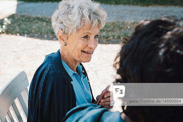 Lächelnde ältere Frau spielt Karten  während sie mit einem männlichen Betreuer im Hinterhof sitzt