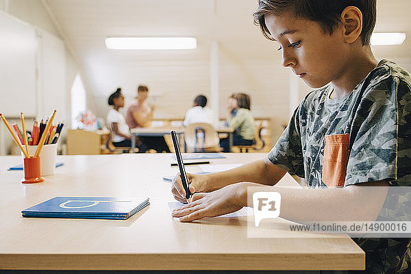 Nahaufnahme eines Jungen  der sich konzentriert  während er am Tisch auf Papier schreibt