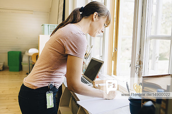 Confident teacher writing on paper while standing by window at classroom in school