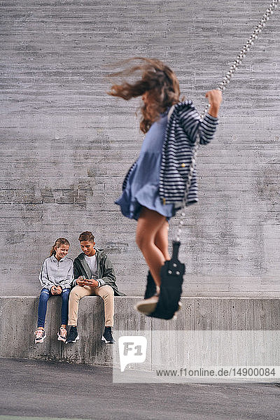 Geschwister in voller Länge beim Mobiltelefonieren  während ein Mädchen auf einer Schaukel auf dem Spielplatz schaukelt