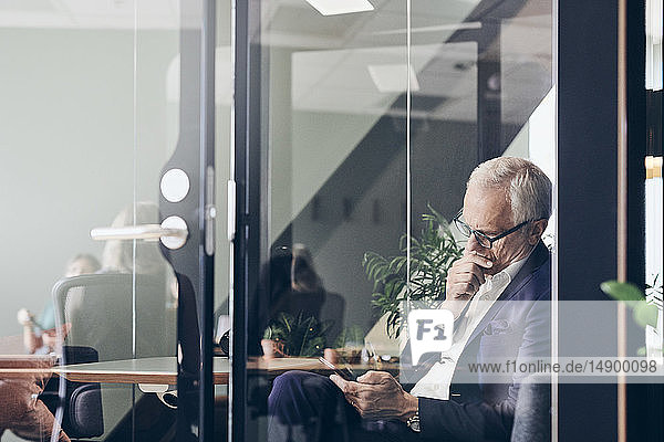 Mature male manager using smart phone seen through glass wall at creative office
