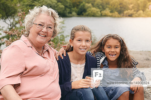 Portrait of smiling granddaughters and grandmother sitting on lakeshore in park
