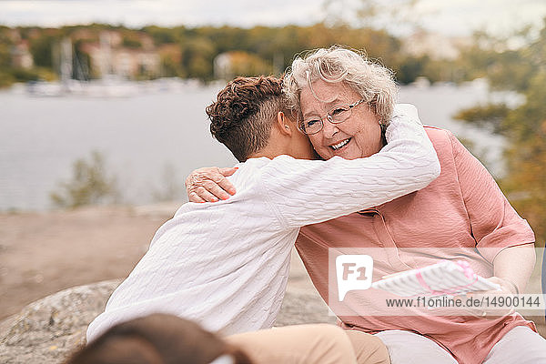 Enkel umarmt lächelnde Großmutter bei der Entgegennahme eines Geschenks am Seeufer im Park während eines Picknicks