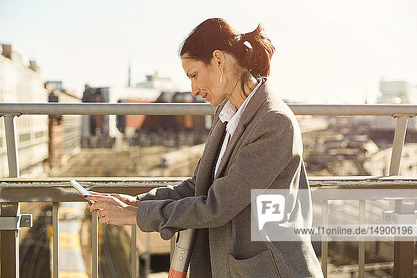 Seitenansicht einer reifen Frau  die ein Smartphone benutzt  während sie mit einem elektrischen Roller auf einer Brücke in der Stadt steht