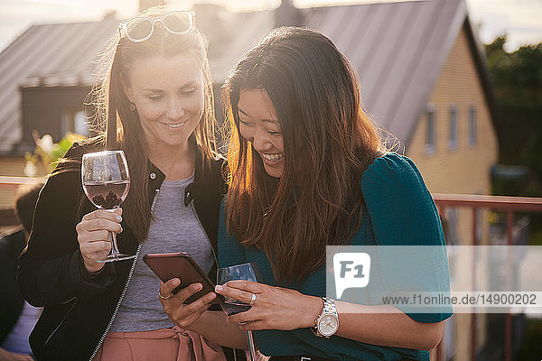 Fröhliche Frau zeigt ihr Handy einer Freundin bei Sonnenuntergang auf der Terrasse