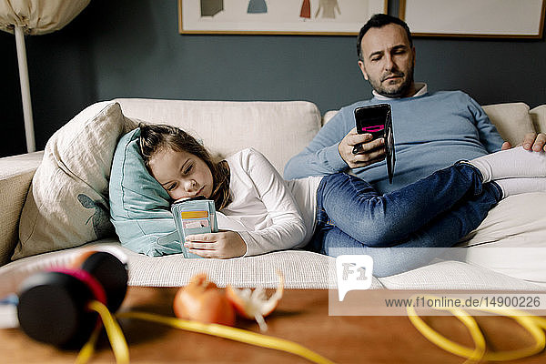 Father and daughter using mobile phones on couch in living room
