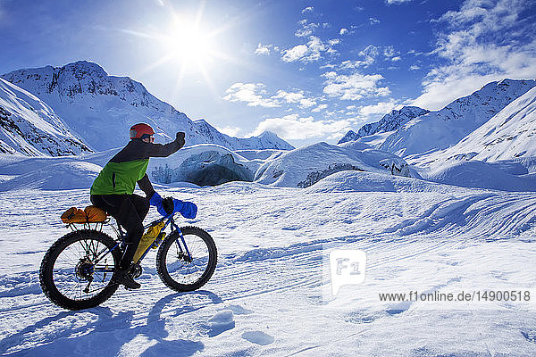 Frau auf dem Fatbike vor dem Skookum-Gletscher  Chugach National Forest  Alaska  an einem sonnigen Wintertag  die Faust pumpt  während sie vorbeifährt  Süd-Zentral-Alaska; Alaska  Vereinigte Staaten von Amerika