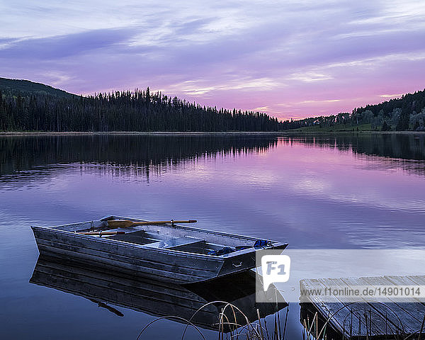 Ein hölzernes Ruderboot liegt neben einem Steg auf einem ruhigen See  in dem sich das Rosa des Sonnenuntergangs spiegelt  Lac Le Jeune Provincial Park; Kamloops  British Columbia  Kanada