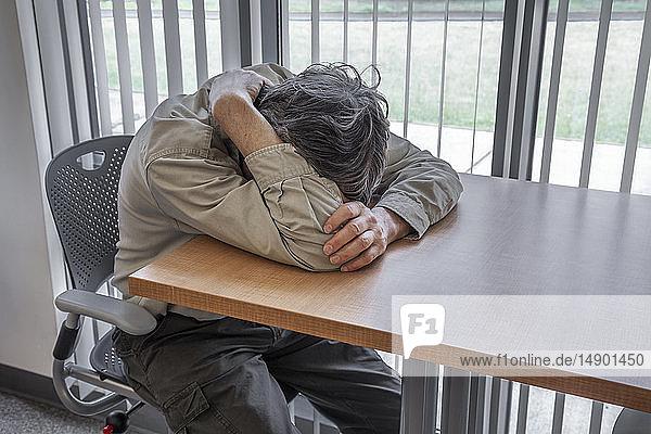 Mann sitzt an einem Tisch in einem Büro vor einem Fenster und hat den Kopf in die Arme gestützt; Connecticut  Vereinigte Staaten von Amerika