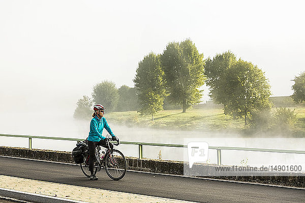 Radfahrerin auf dem Flussweg bei Nebel im Flusstal mit Blick auf die Bäume am Flussufer  nördlich von Remich; Luxemburg