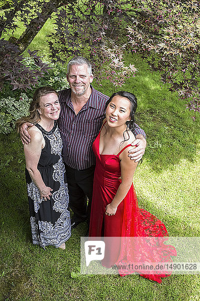 Porträt einer High-School-Absolventin in einem roten Gesellschaftskleid  die mit ihren Eltern in einem Hof steht; Chilliwack  British Columbia  Kanada