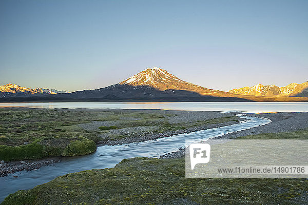 Ein Bach führt den Blick auf einen hoch gelegenen See. Ein Vulkan und schneebedeckte Berge in der Ferne werden vom Sonnenaufgang vor einem rein blauen Himmel beleuchtet; Mendoza  Argentinien