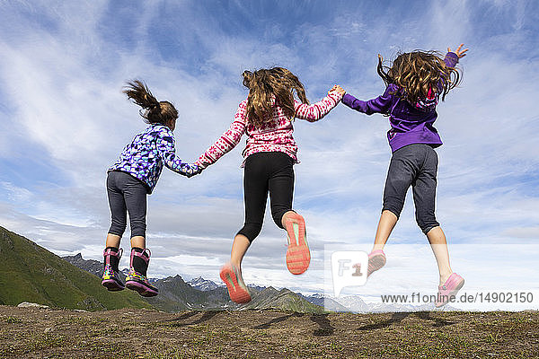 Three young sisters jumping and holding hands in the Talkeetna Mountains  Hatcher’s Pass; Alaska  United States of America