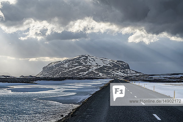 Straße  die in die dramatische Landschaft Islands führt  während die Sonne durch die Wolken scheint und ein schönes Bild abgibt; Island