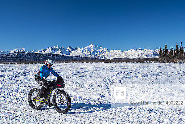 A man riding a fatbike on the Chulitna Bluff Trail on a sunny winter day. The Alaska Range and 20 230' Mount Denali (McKinley) in seen in the background  South-central Alaska; Alaska  United States of America