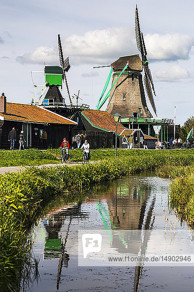 Historische hölzerne Windmühlen  die sich in einer kleinen Gracht mit Radweg und Radfahrern spiegeln; Zaandam  Niederlande