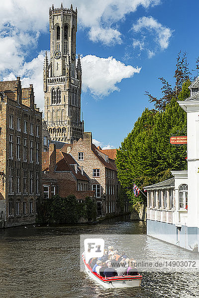 Small touring boat with people in a canal with large gothic church tower in the background; Bruges  Belgium