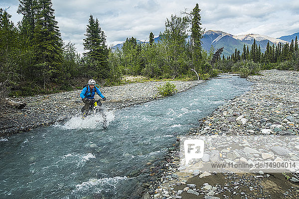 A man fat biking across a stream in Wrangell-St. Elias National Park and Preserve on a cloudy summer day in South-central Alaska; Alaska  United States of America