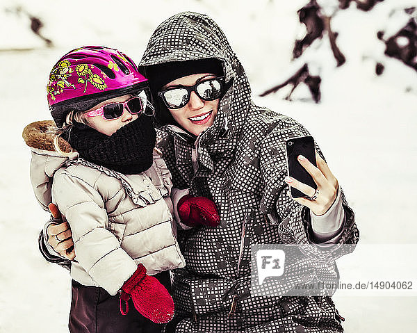 A Mother And Daughter Dressed In Winter Coats  Hats And Sunglasses Taking A Self-Portrait With A Smart Phone While Tobogganing on Vacation; Fairmont Hot Springs  British Columbia  Canada
