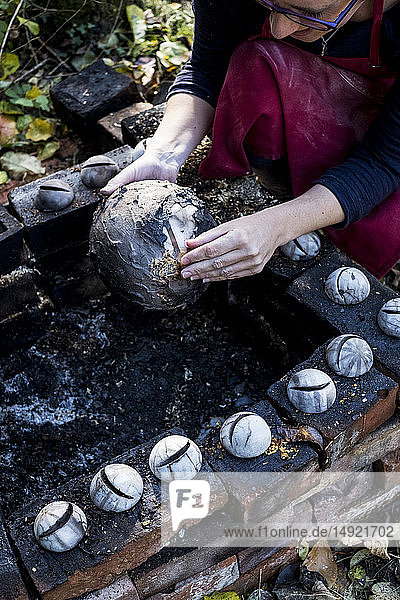 Hochwinkel-Nahaufnahme eines Keramikkünstlers  der neben einer Rauchfeuergrube im Freien sitzt und an einer Vase arbeitet.