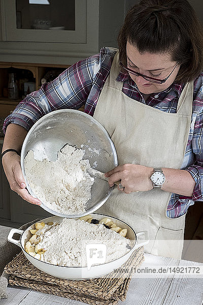 Frau mit Schürze  die in einer Küche steht und frische Streuselmischung auf eine mit Obst gefüllte Kuchenform gießt.