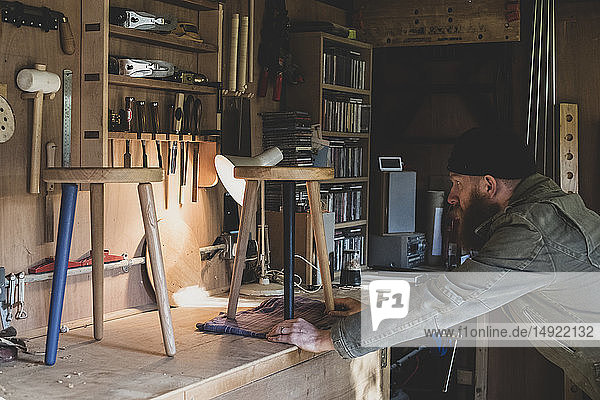 Bearded man wearing black beanie standing at workbench in workshop  examining wooden stool.