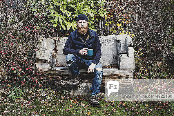 Bärtiger Mann mit schwarzer Mütze auf Holzbank im Garten sitzend  blaue Tasse in der Hand  in die Kamera blickend.