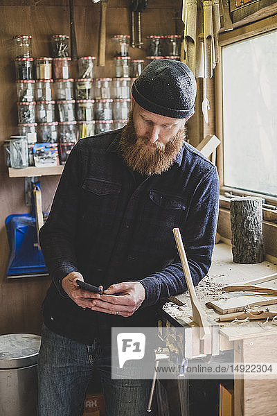 Bearded man wearing black beanie standing next to workbench in workshop  checking his mobile phone.