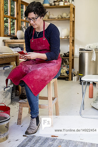 Frau mit roter Schürze sitzt auf einem Hocker in ihrer Keramikwerkstatt und überprüft ihr Mobiltelefon.