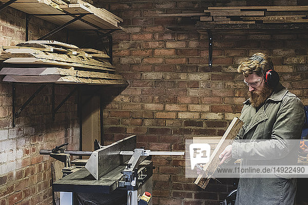 Bärtiger Mann steht in der Werkstatt  trägt Gehörschutz und arbeitet an einem Stück Holz.