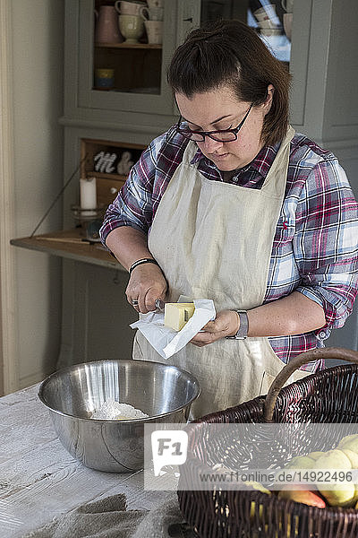 Woman wearing apron standing in a kitchen cutting butter pieces into a metal bowl filled with flour.