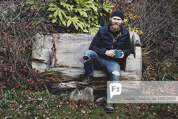 Bearded man wearing black beanie sitting on wooden bench in garden  holding blue mug  looking at camera.