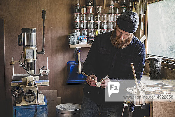 Bearded man wearing black beanie standing at workbench in workshop  examining piece of wood.