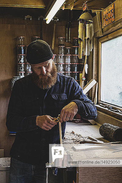 Bärtiger Mann mit schwarzer Mütze  der an der Werkbank in der Werkstatt steht und an einem Stück Holz arbeitet.