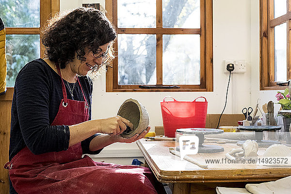 Frau mit roter Schürze sitzt in ihrer Keramikwerkstatt und arbeitet an einer kleinen Tonschüssel.