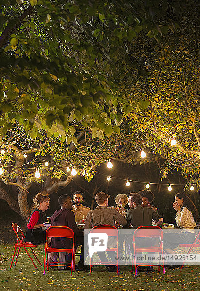 Freunde genießen das Abendessen Gartenparty unter Bäumen mit Lichterketten