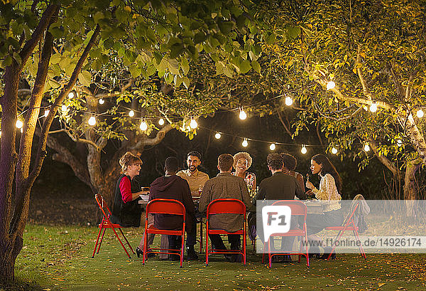 Freunde genießen das Abendessen Gartenparty unter Bäumen mit Lichterketten