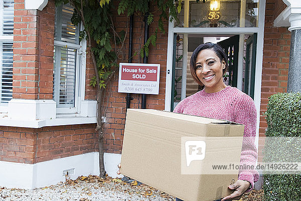 Porträt einer lächelnden  selbstbewussten Frau  die in ein neues Haus einzieht und einen Karton vor dem Haus hält