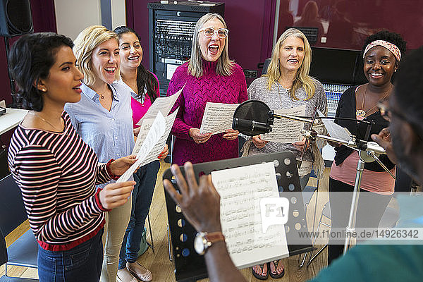 Male conductor leading womens choir with sheet music singing in music recording studio