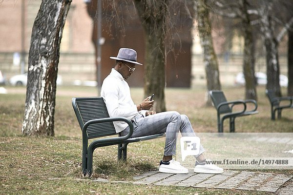 Stylish man sitting alone on bench in park  resting  using phone  looking aside  in city Munich  Germany.
