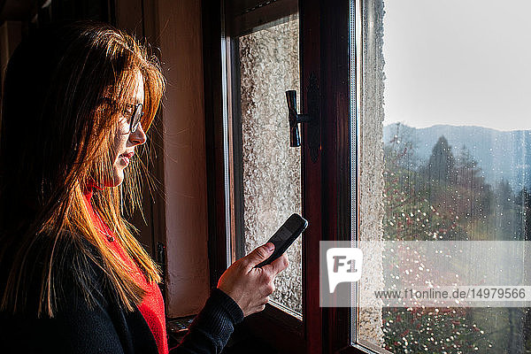 Junge Frau mit langen roten Haaren betrachtet Smartphone am Fenster